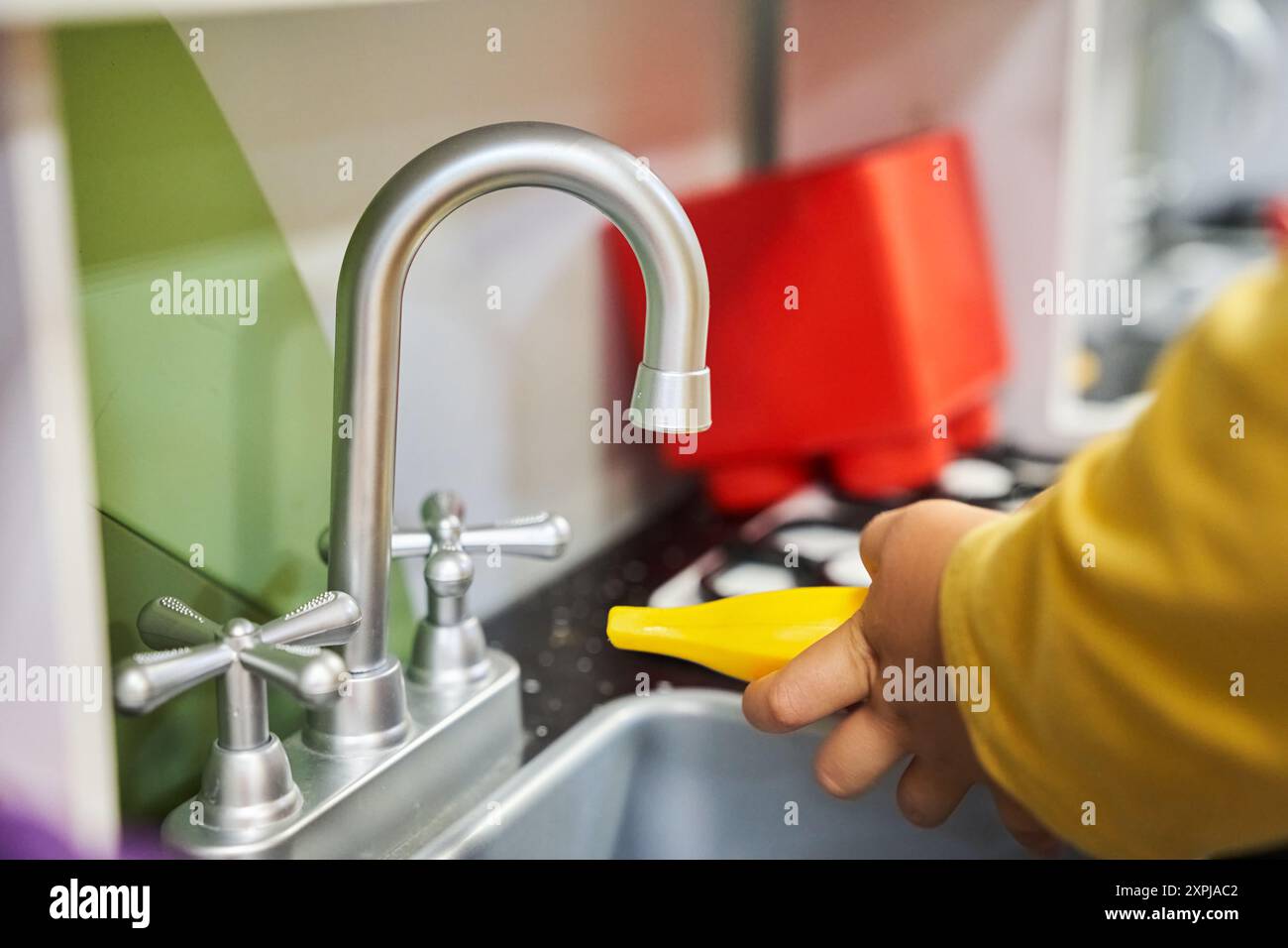 Un bambino irriconoscibile sta giocando nella stanza dei bambini. Le mani del bambino tengono una banana giocattolo vicino al lavandino della cucina giocattolo. Con spazio per copiare. Foto di alta qualità Foto Stock