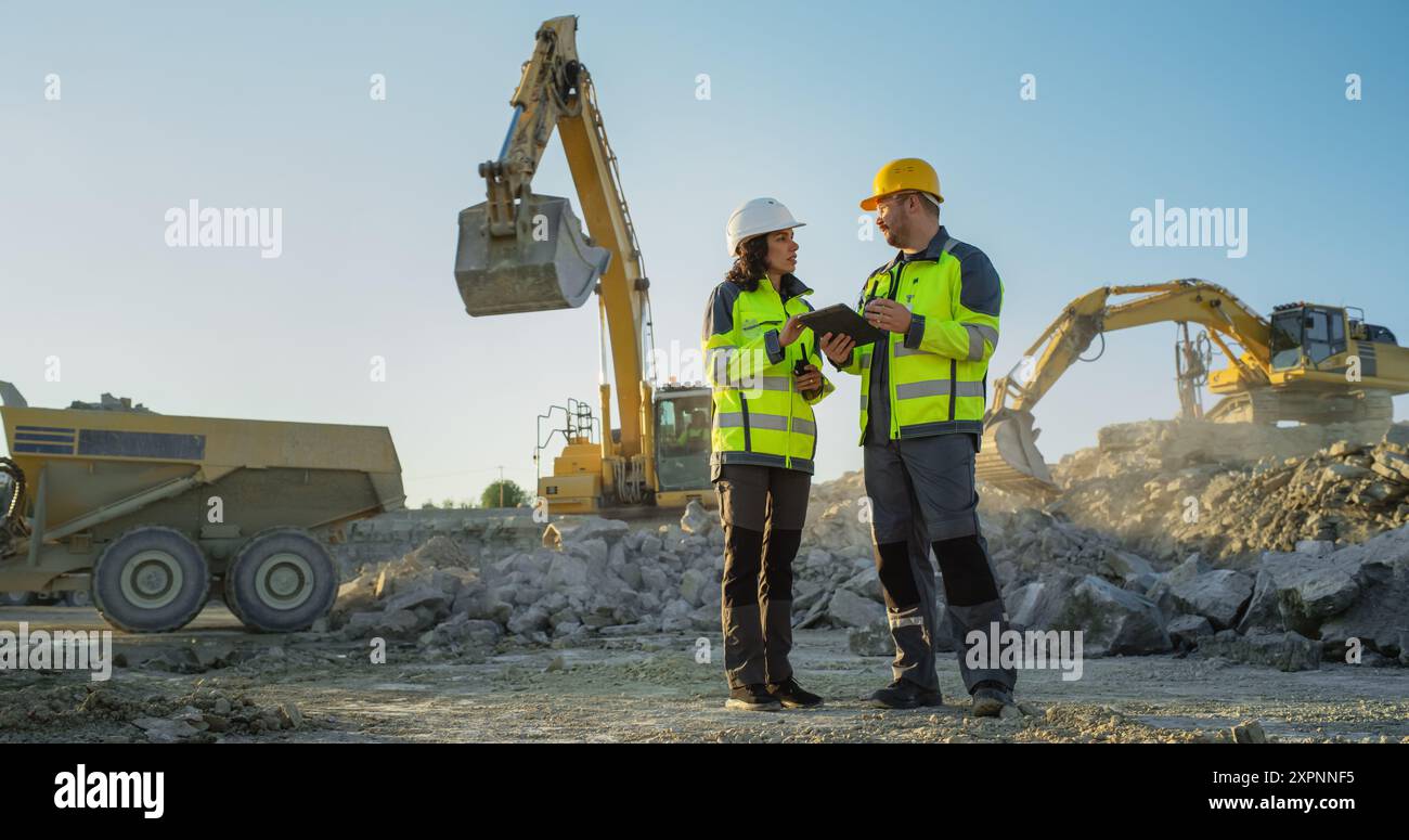 Ispettore femminile ispanico che parla con il responsabile dello sviluppo del territorio maschile caucasico con tablet on Construction Site of Real Estate Project. Escavatori industriali in preparazione per la posa di fondamenta per l'edilizia. Foto Stock