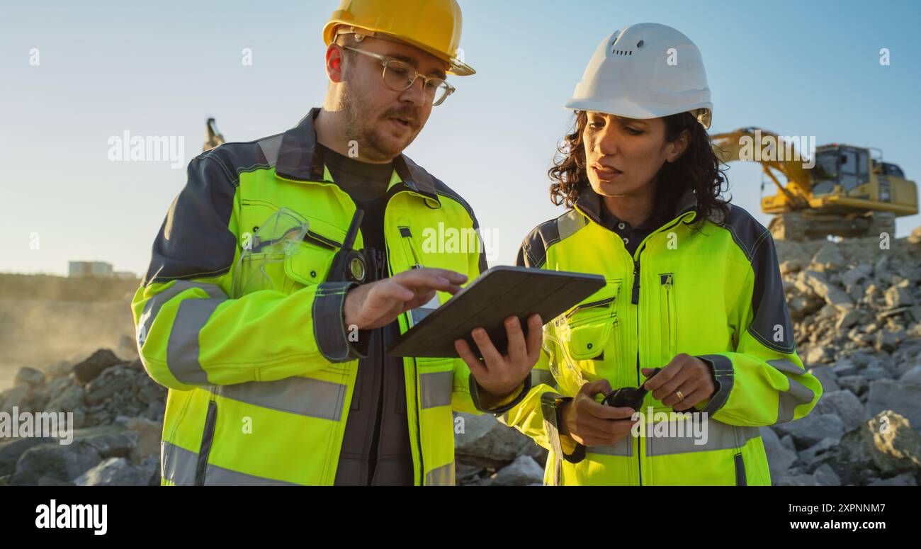 Ingegnere civile uomo caucasico che parla con ispettore donna ispanico e utilizza tablet sul sito di costruzione di un nuovo edificio residenziale. Sviluppatori immobiliari che discutono di affari, escavatori che lavorano. Foto Stock