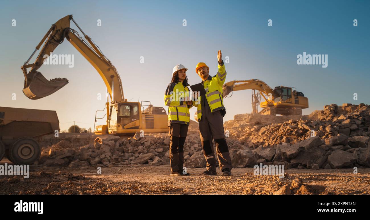 Ispettore femminile ispanico che parla con il responsabile dello sviluppo del territorio maschile caucasico con tablet on Construction Site of Real Estate Project. Escavatori in preparazione per la posa di fondamenta edili. Caldo giorno di sole Foto Stock