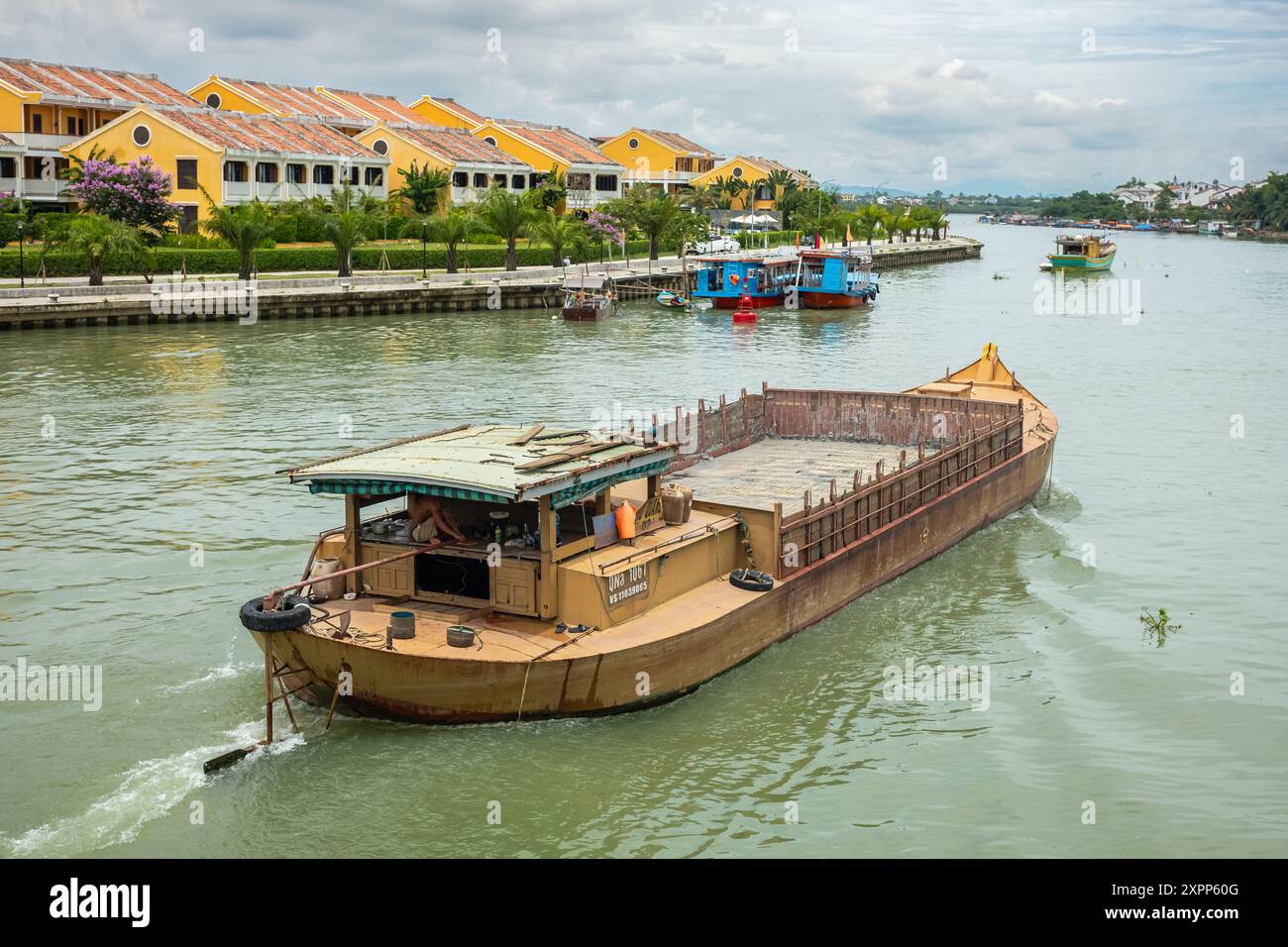 La chiatta viaggia sul fiume. Una chiatta galleggiante sul fiume Thu Bon a Hoi An Vietnam. Città antica, case colorate, container di spedizione su chiatta fluviale. Foto Stock