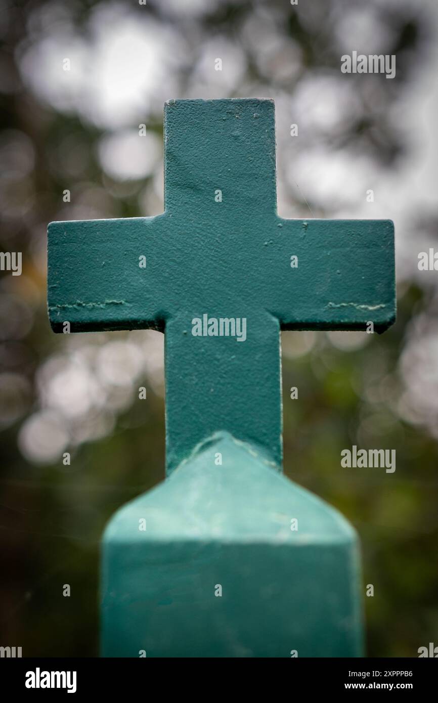 Foto ravvicinata di una croce cristiana su una recinzione verde ferro del cimitero su uno sfondo sfocato in un ambiente naturale con effetto bokeh Foto Stock