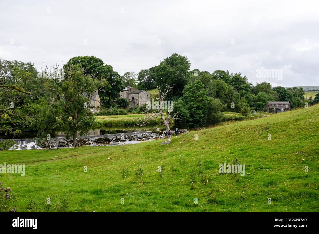 Grassington Yorkshire Regno Unito - 27 luglio 2024. Tranquillo scenario di campagna con una storica casa in pietra in riva a un fiume circondato da vegetazione lussureggiante. Foto Stock