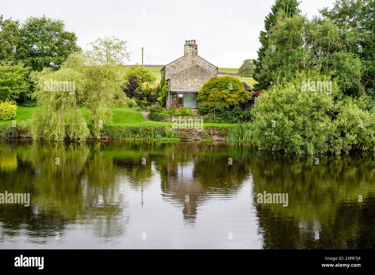 Grassington Yorkshire Regno Unito - 27 luglio 2024. Pittoresco cottage di campagna con vegetazione lussureggiante che si riflette su un fiume tranquillo. Foto Stock