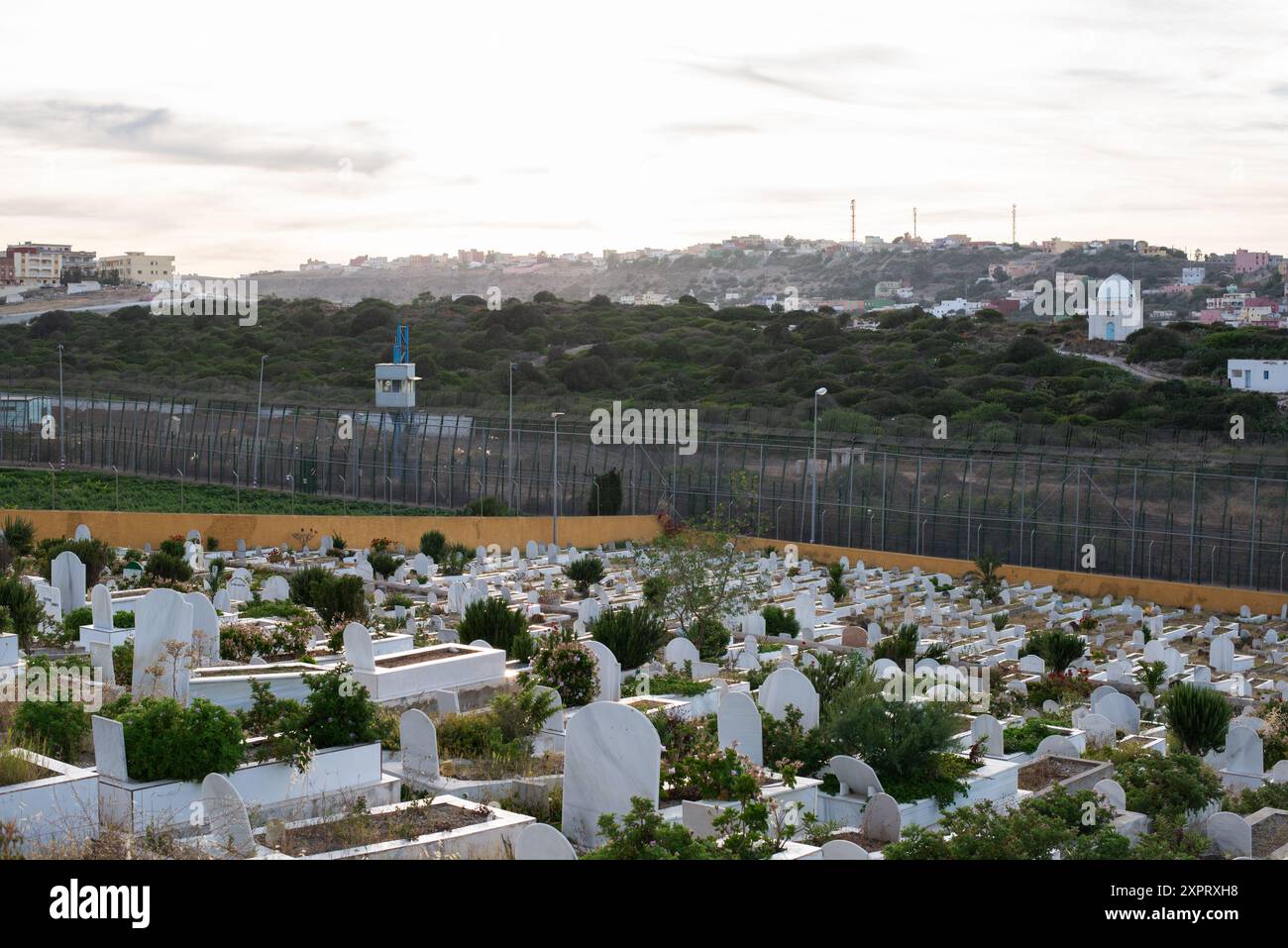 Immagine che raffigura una recinzione ad alta sicurezza con fili alti sei metri e sistemi di sorveglianza come metafora per divisione e barriere. Il cimitero solenne in primo piano aggiunge profondità al concetto di separazione. Foto Stock