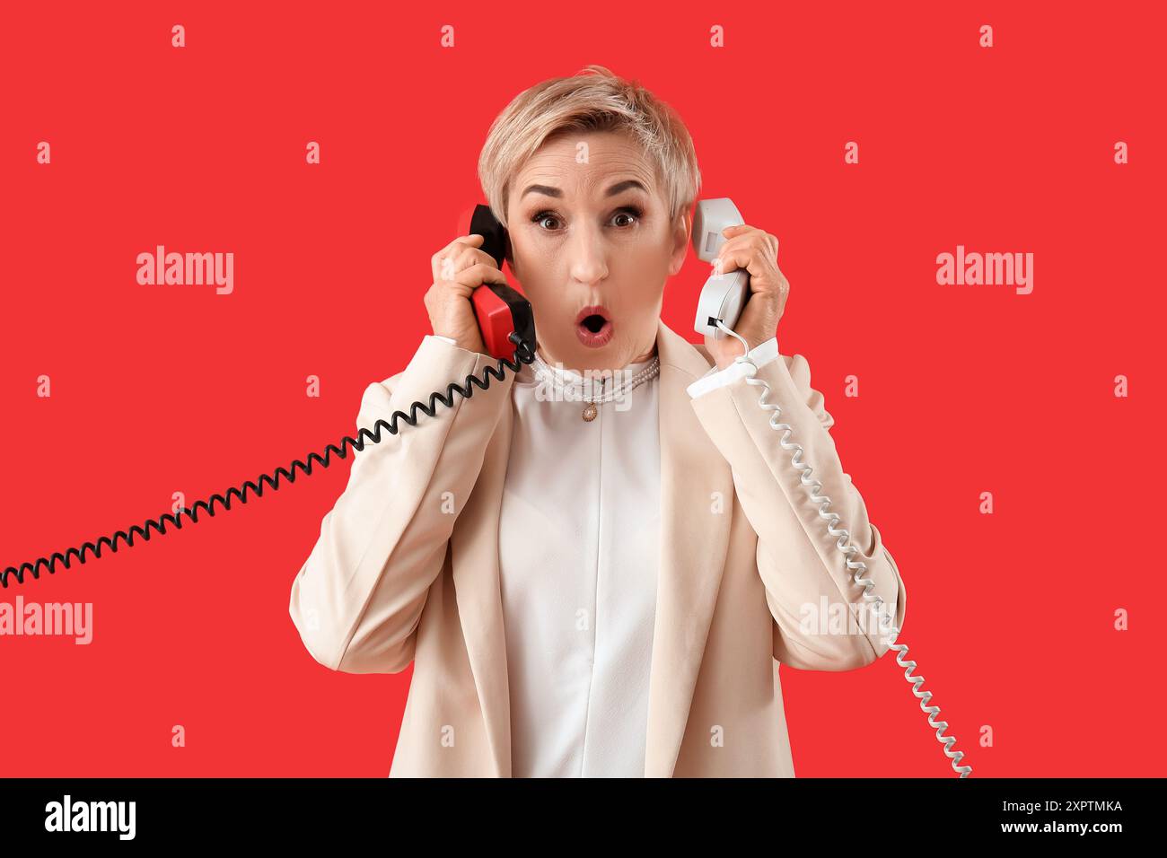 Donna matura scioccata con due ricevitori telefonici su sfondo rosso Foto Stock