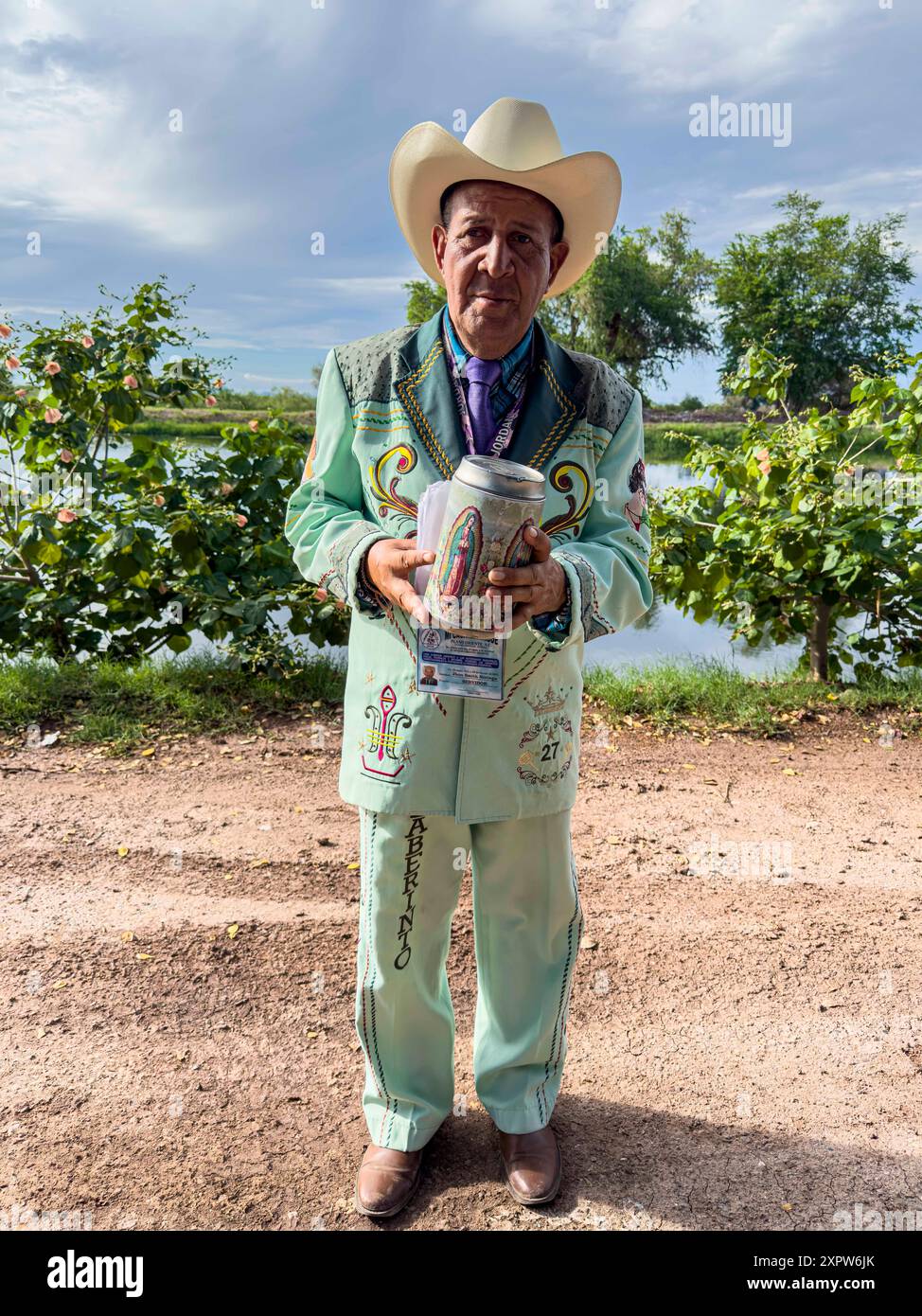 Jhon Smith Noriega posa per un ritratto accanto al canale superiore a Laguna del Nainari nella città di Obregón, Sonora, Messico, il 14 luglio 2024. Jhon indossa un costume del gruppo musicale popolare o regionale la banda chiamato lLaberinto mentre chiede sostegno finanziario con una barca con l'immagine della Vergine di Guadalupe per "mi Casa Albergue" (foto di Luis Gutiérrerez / Norte foto) Jhon Smith Noriega posa para un retrato junto al Canal alto en la Laguna del Nainari en ciudad Obregón Sonora México el 14 julio 2024. Jhon viste de traje de la agrupación de músical Popular o Regional de la banda Foto Stock