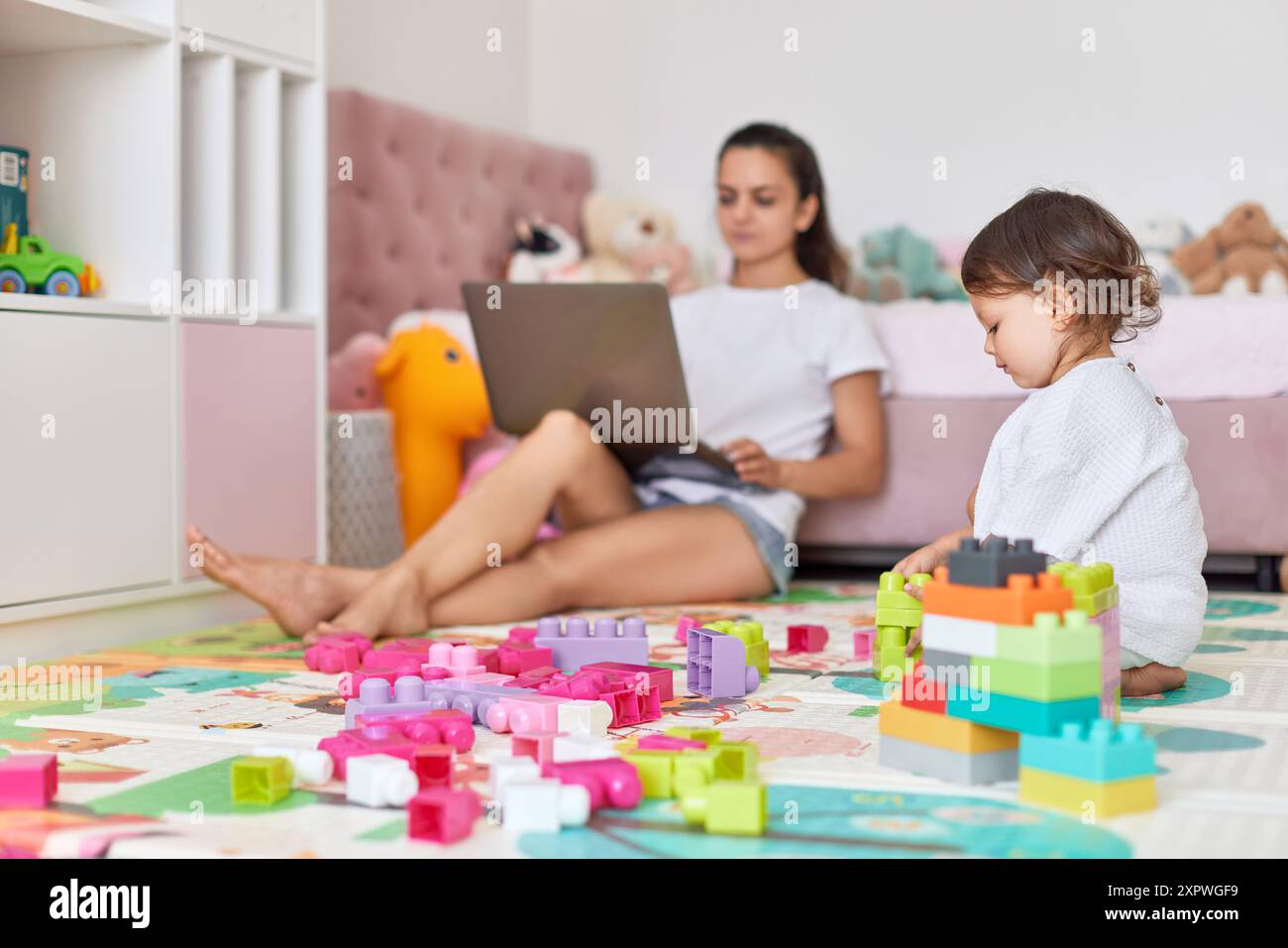 una bambina carina che gioca con i blocchi di giocattoli e una madre che lavora seduta sul pavimento con un computer portatile in camera Foto Stock