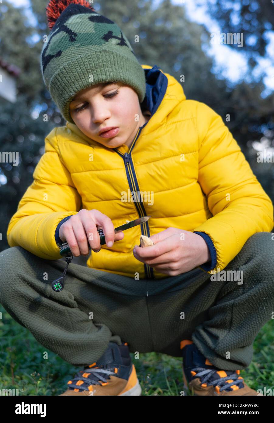 Ragazzo caucasico di 8 anni concentrato che affina un bastone con un rasoio in natura nei vestiti invernali Foto Stock