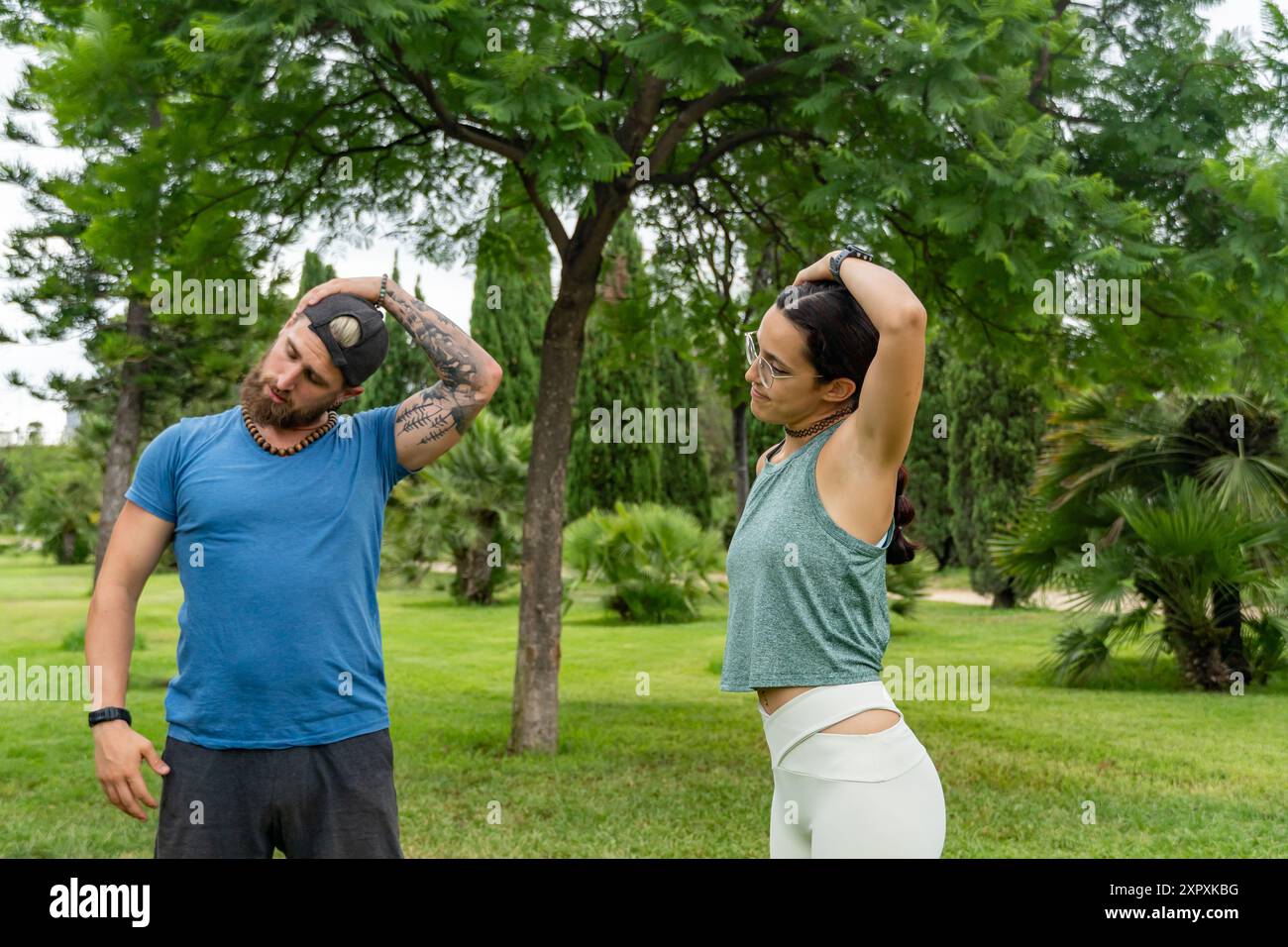 Un giovane uomo con la barba rossa e una giovane donna si stanno concentrando sul collo che si allunga insieme in un parco all'aperto. Allenamenti di coppia, routine di stretching e a Foto Stock