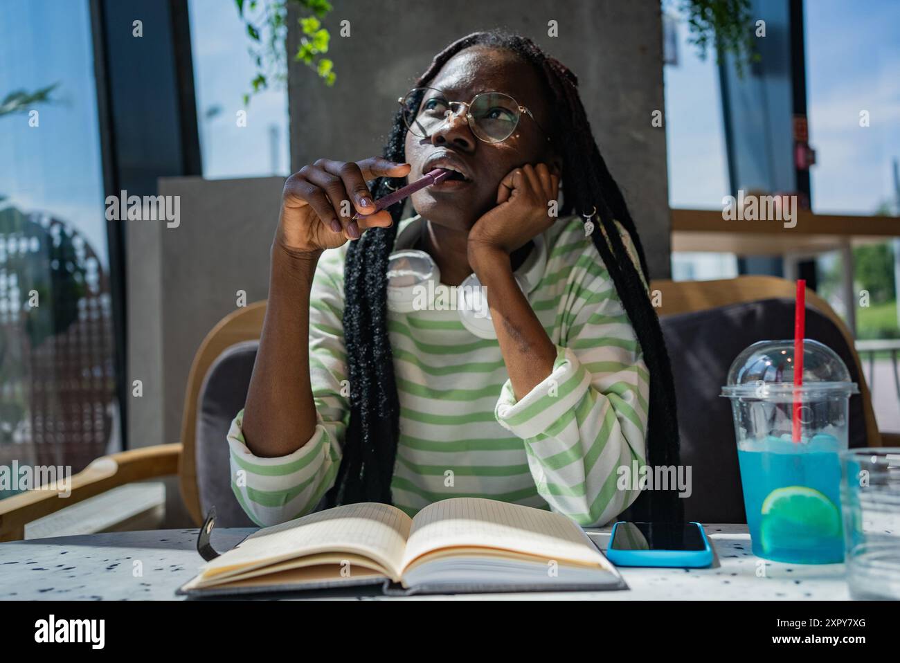 Giovane donna afro-americana con trecce e occhiali che contemplano mentre scrive in un taccuino in un bar, con cuffie intorno al collo e un drink nelle vicinanze. Foto Stock