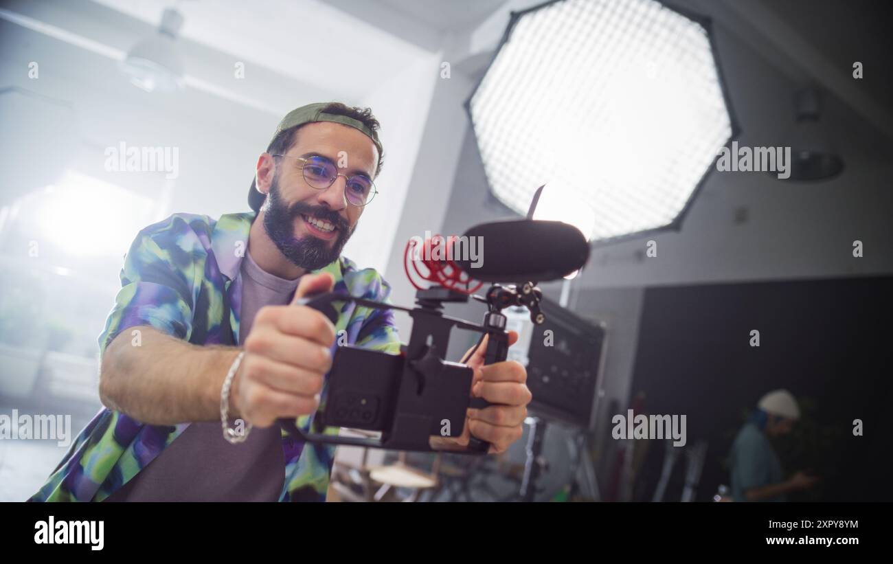 Giovane videografo caucasico maschio con barba, indossa camicia e berretto colorati, utilizza con entusiasmo la fotocamera in ambiente Studio, catturando contenuti per la pubblicità o la produzione cinematografica. Foto Stock