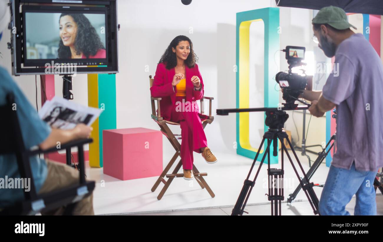La giovane donna ispanica in Un vivace vestito rosso parla animatamente su Una sedia del regista, circondata da scenografie colorate, con Un direttore della fotografia maschile che registra per Una vivace trasmissione televisiva o web. Foto Stock