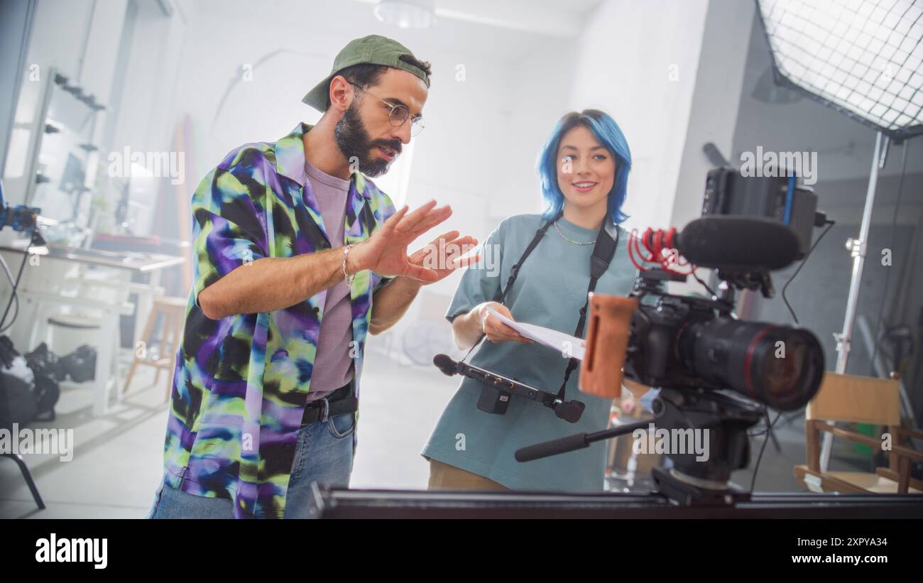 Giovane regista latino con barba e donna caucasica con capelli blu discutono su Una sceneggiatura in Uno studio cinematografico ben attrezzato, circondato da macchine fotografiche professionali e apparecchiature di illuminazione. Foto Stock