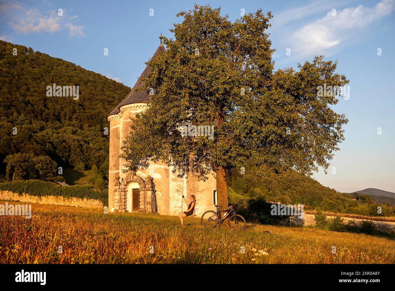 Turista donna, ciclista in gita in bicicletta, siede su un'altalena sotto un bellissimo albero con la Torre del Diavolo nel villaggio di Soteska, regione di Dolenjska, Slovenia Foto Stock