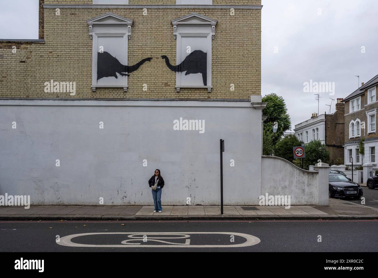 Londra, Regno Unito. 8 agosto 2024. Una donna sotto un'opera d'arte di Banksy raffigurante un paio di elefanti sul lato di un edificio vicino a Chelsea. Nell'ultima settimana, nella capitale sono apparse quotidianamente nuove opere dell'artista con un tema animale. Finora il sentiero comprende una capra di montagna, due elefanti e tre scimmie. Una quarta opera, raffigurante un lupo ulululante dipinto su una parabola satellitare a Peckham, è già stata rubata poco dopo essere stata rivelata oggi. Crediti: Stephen Chung / Alamy Live News Foto Stock