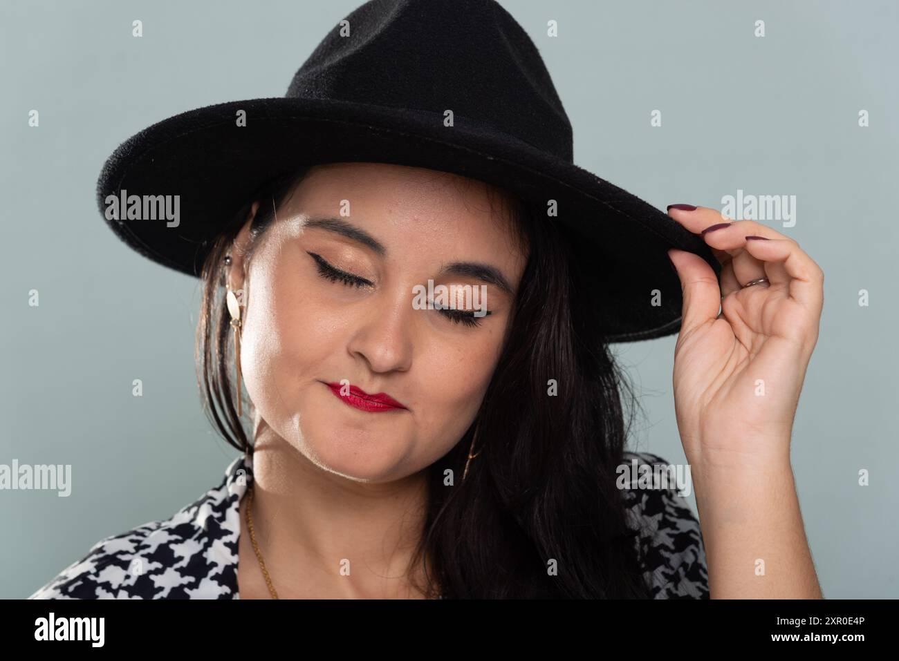 Ritratto ravvicinato di una bella donna sorridente con i capelli neri che indossa un cappello nero in posa per la foto in un abito bianco e nero. Isolato su verde chiaro b Foto Stock