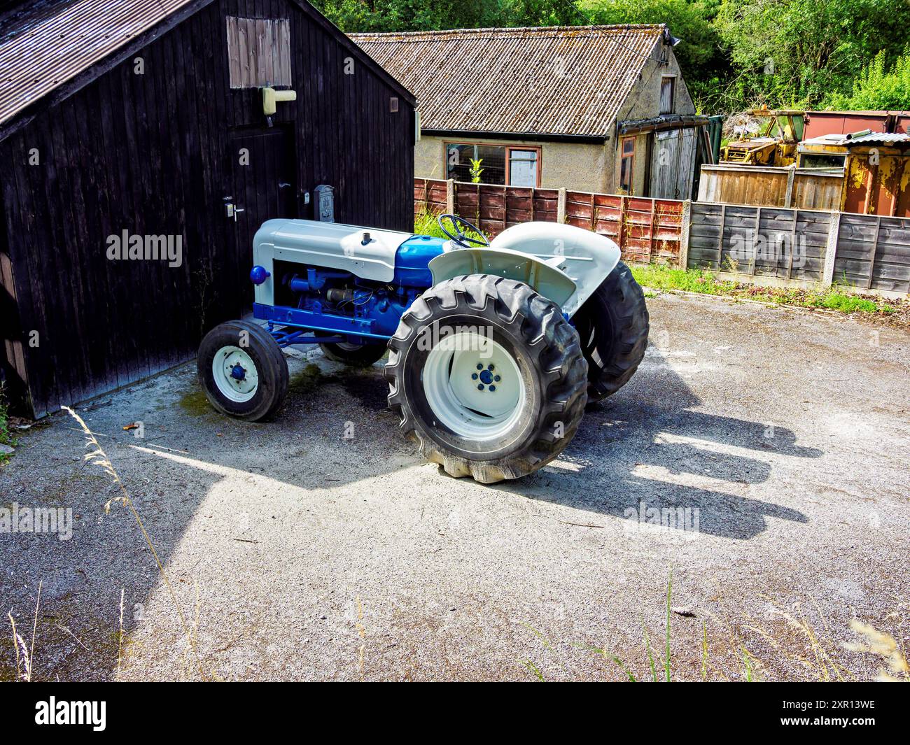 Trattore bianco e blu d'epoca parcheggiato vicino a un rustico capannone di legno in una giornata di sole. Foto Stock