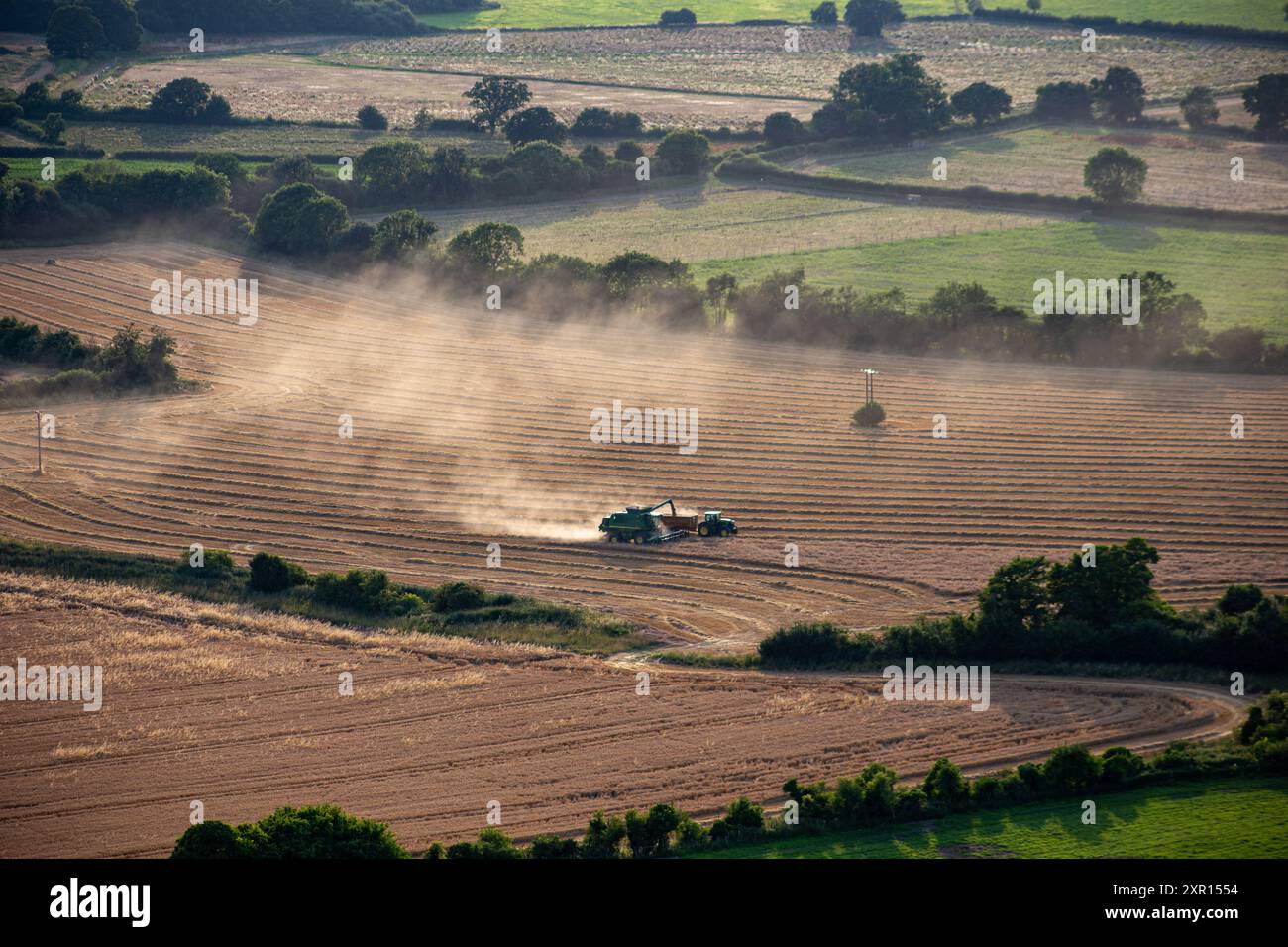 Vista aerea di un trattore che lavora su un terreno agricolo, creando nuvole di polvere, circondato da campi verdi e alberi in un paesaggio rurale. Foto Stock