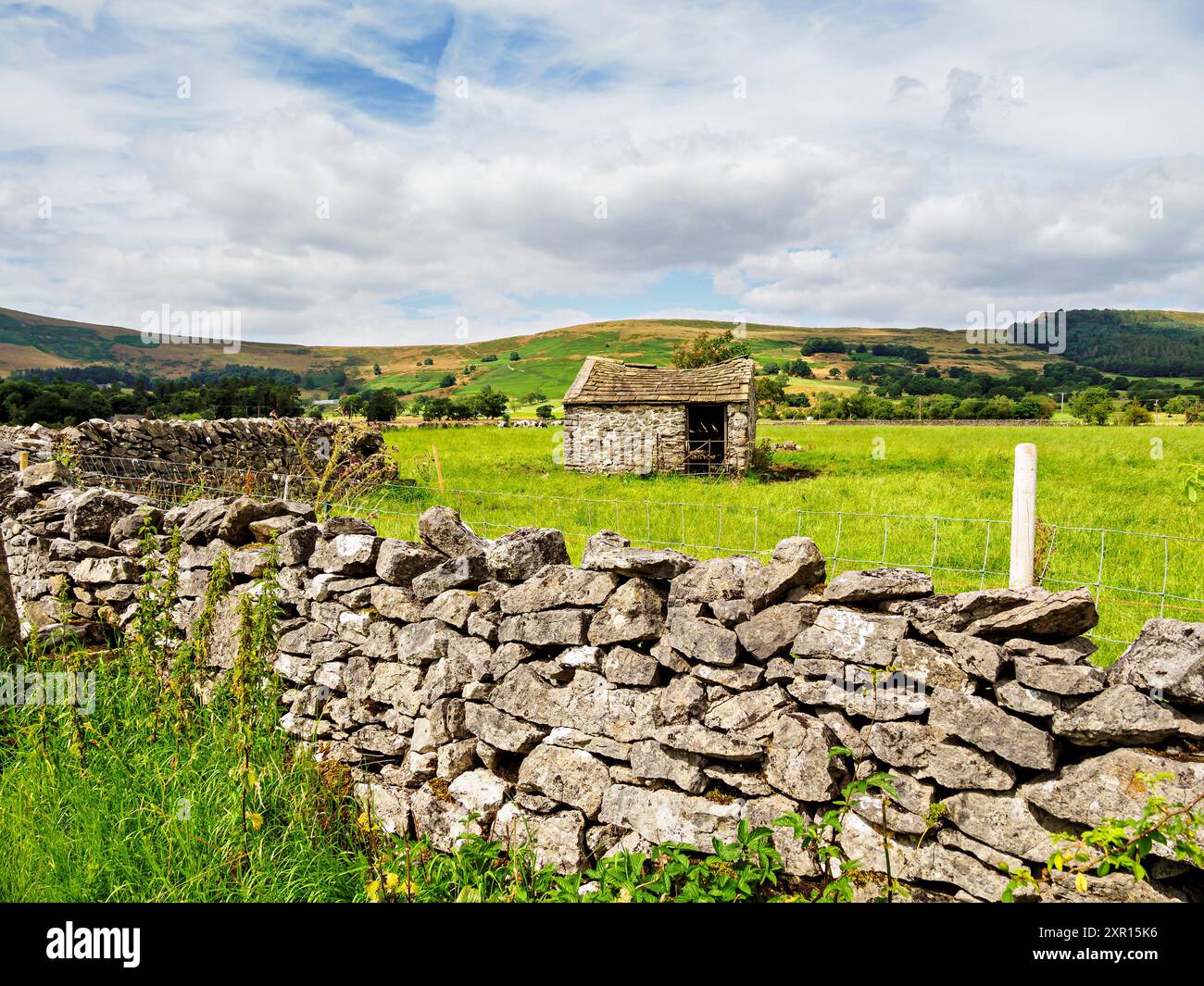 Un affascinante capannone di pietra circondato da lussureggianti campi verdi e da un muro di pietra intemprata in una giornata luminosa e parzialmente nuvolosa in campagna. Foto Stock