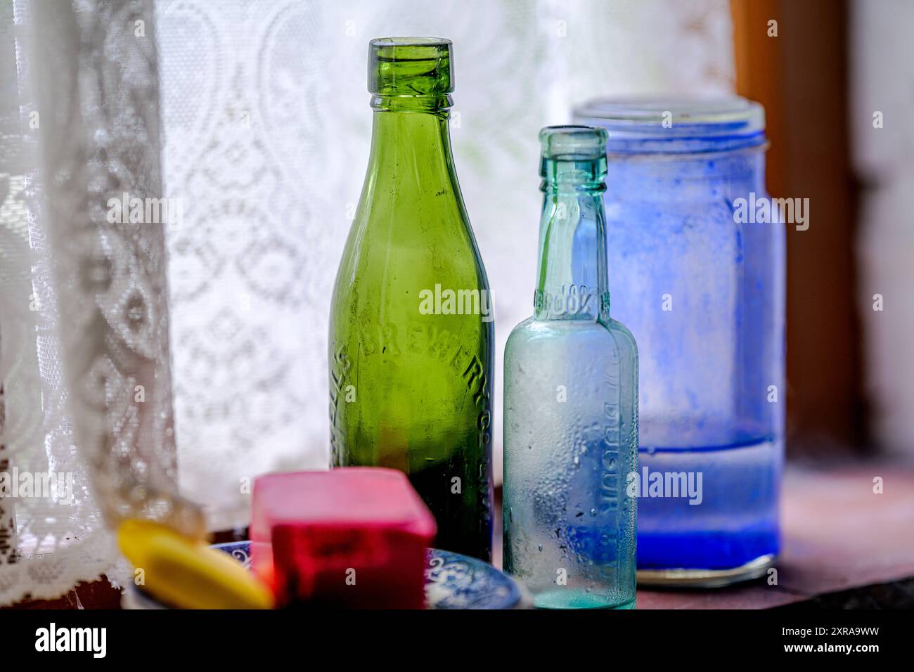 vecchi oggetti di vetro illuminati dalla luce di una vecchia finestra con tende a rete Foto Stock