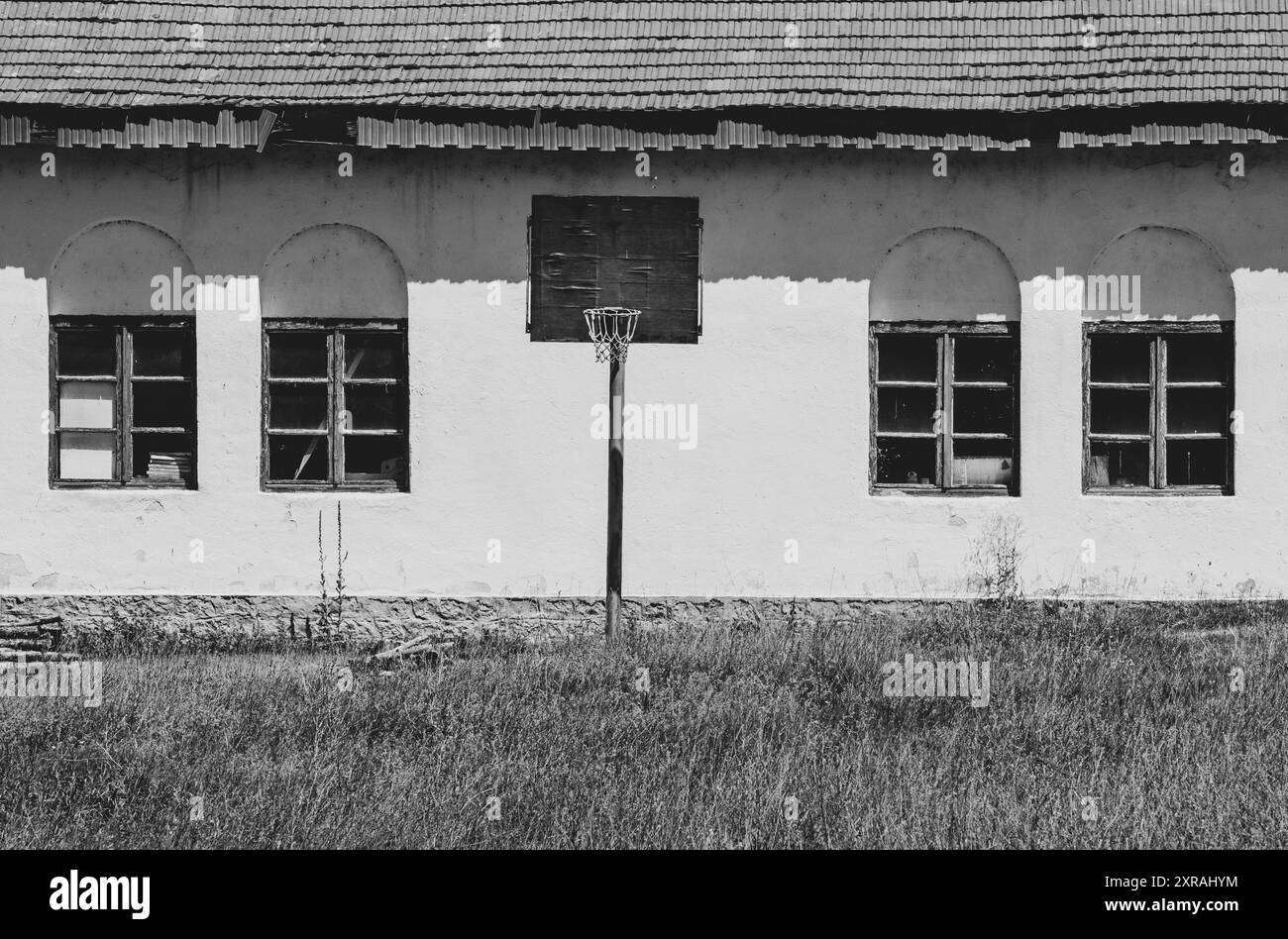 Vecchio edificio abbandonato, cortile scolastico con palo da basket e rete. Cornici Windows. Simmetria. Bianco e nero. Monocromatico. Foto Stock