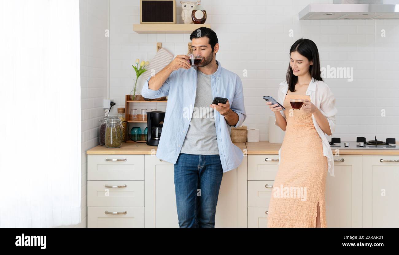 Giovane coppia che beve caffè caldo in cucina con lo smartphone. Donna latina sorridente guardando il fidanzato mentre si siede sul bancone della cucina Foto Stock
