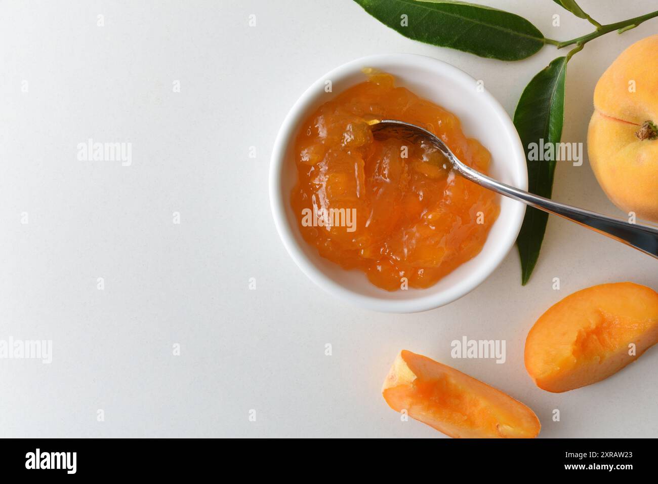 Dettaglio di un piccolo piatto con marmellata di pesca su un banco bianco con frutta e foglie intorno ad esso. Vista dall'alto. Foto Stock