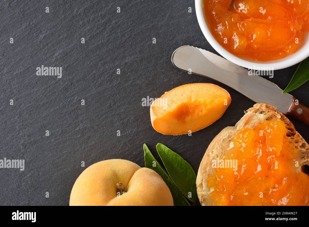 Colazione macro con marmellata di pesca su una fetta di pane e su un recipiente di ceramica bianca con frutta intorno su un piatto di ardesia. Vista dall'alto. Foto Stock