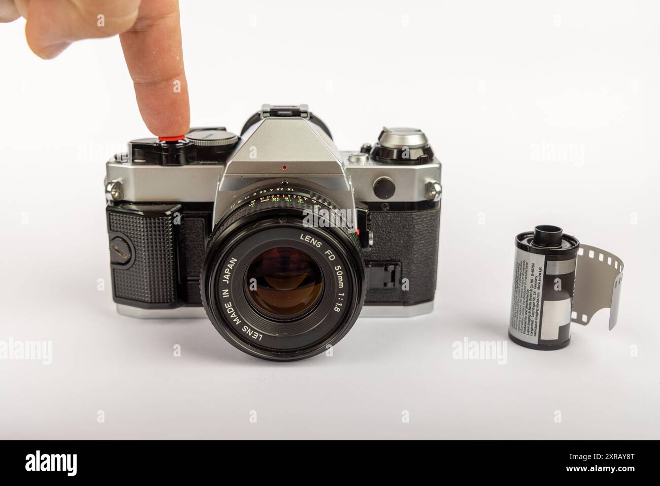 Una vecchia fotocamera e rotoli di pellicola da 35 mm su sfondo bianco. La fotocamera è centrata, con un dito che preme il pulsante dell'otturatore Foto Stock