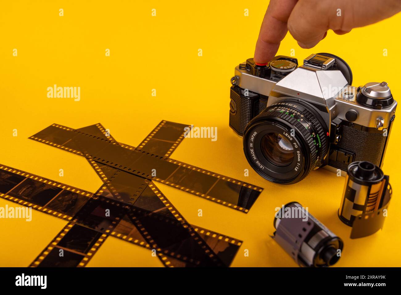 Un dito preme l'otturatore su una fotocamera antica, circondata da rotoli di pellicola sviluppati da 35 mm. La scena è evidenziata da uno sfondo giallo. Foto Stock