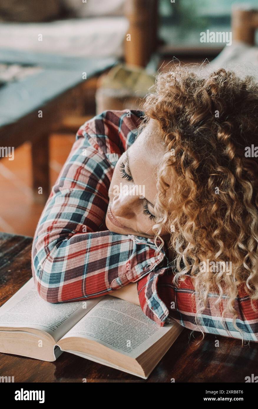 Donna carina che dorme su un libro aperto da sola a casa. Donne stanche che dormono sul tavolo. Concetto di stile di vita degli studenti. Una bella bionda lunga e riccia che riposa nel soggiorno. Abitacolo accogliente Foto Stock