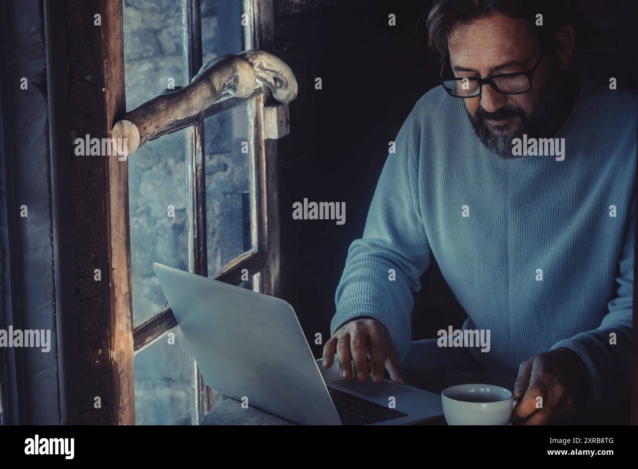 Bell'uomo maturo che lavora con un computer portatile al buio all'interno di un'accogliente cabina di legno. Concetto di lavoro intelligente e attività di svago tecnologica al chiuso con persone di sesso maschile che scrivono Foto Stock