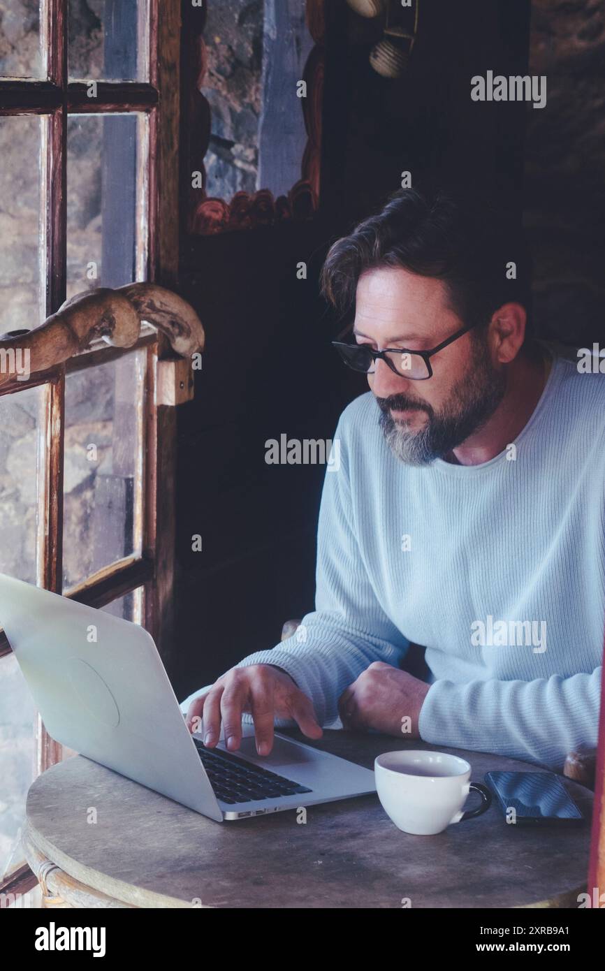 Uomo maturo al lavoro su computer portatile seduto al tavolo all'interno di una casa confortevole cabina con finestre in legno - digitale intelligente lavoro adulti concetto stile di vita Foto Stock