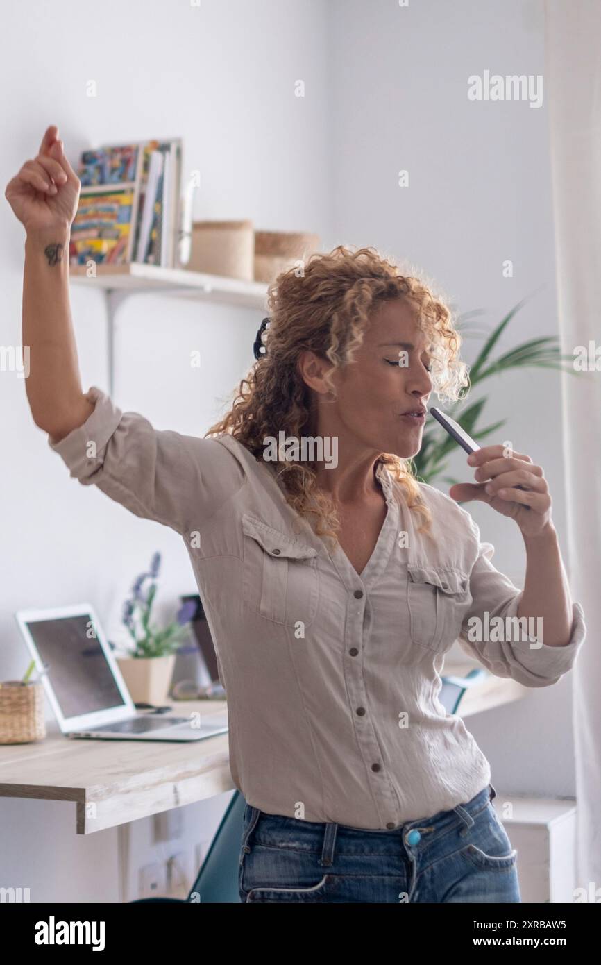 Una donna a casa da sola che canta al telefono e balla per la felicità. Benessere e stile di vita indipendente: Attività ricreative all'interno della porta. Una bella ragazza che si diverte in salotto Foto Stock