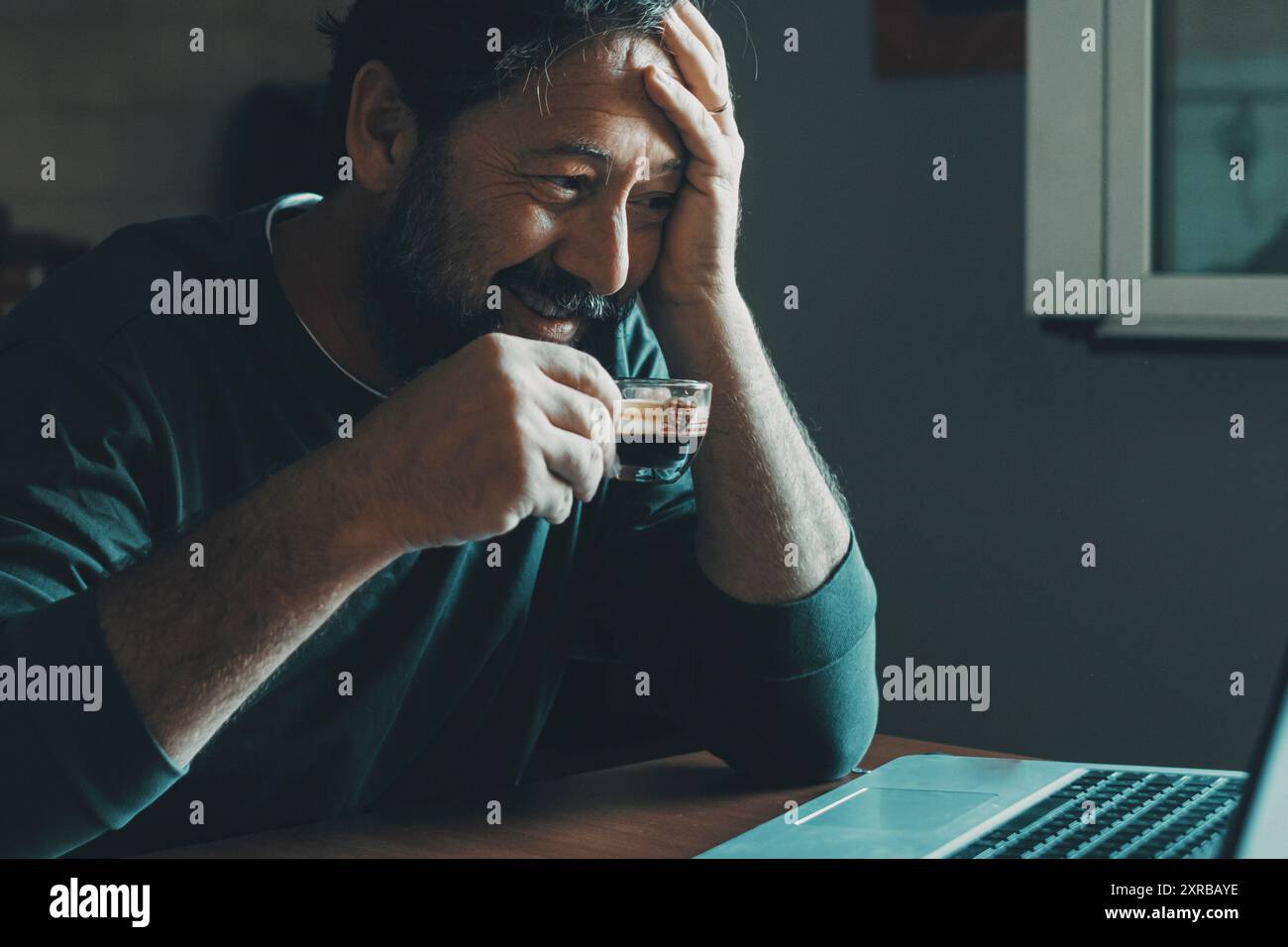 L'uomo da solo a casa usa un computer portatile sorridente e beve caffè espresso in cucina. Lavoro mattutino di mattina presto e tecnologia Internet concetto di attività per il tempo libero al coperto stile di vita. Persone moderne che usano il computer Foto Stock