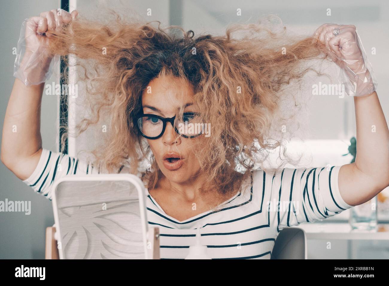 Una giovane donna a casa sembra in cattive condizioni dei suoi capelli pronta all'uso e applica un trattamento di bellezza da sola seduta al tavolo. Acconciatura self-care all'interno degli appartamenti. Persone indipendenti Foto Stock
