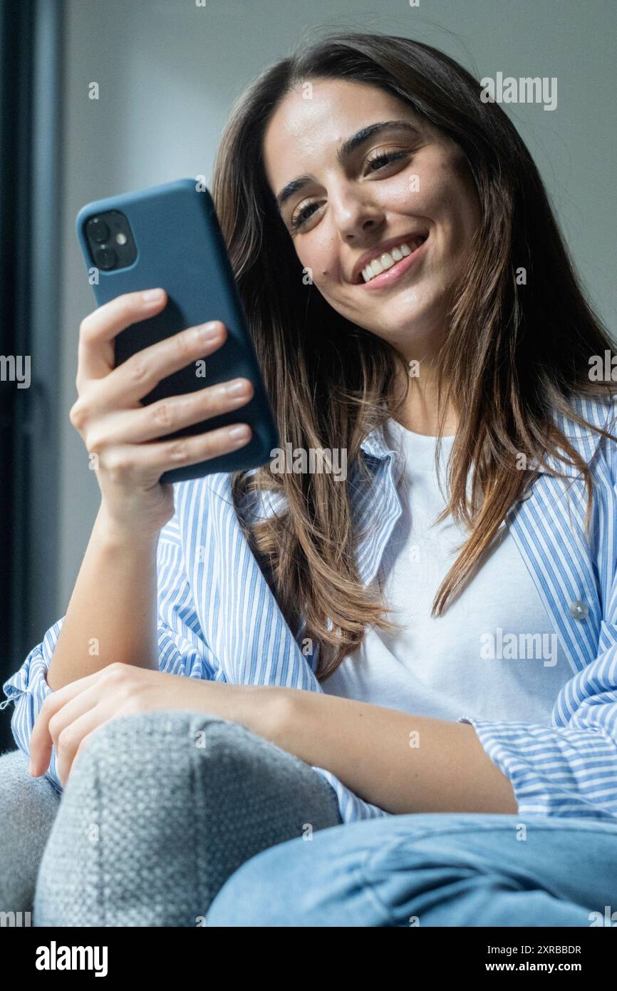 Donna felice e piacevole, millenaria, che si rilassa sul comodo divano, tenendo lo smartphone nelle mani. Sorridente giovane donna che chiacchiera nei social network, guardando f Foto Stock