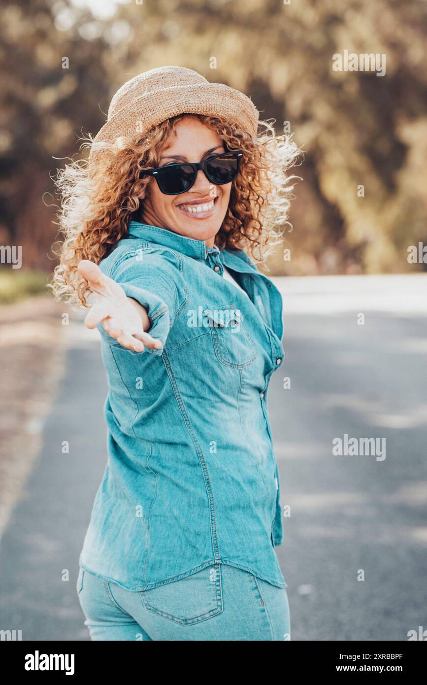 Ritratto di una splendida modella bianca con capelli biondi ricci che esprimono energia in una buona giornata in Europa. Bella donna riccia che sorride e cammina con gli alberi dietro di lei. Foto Stock