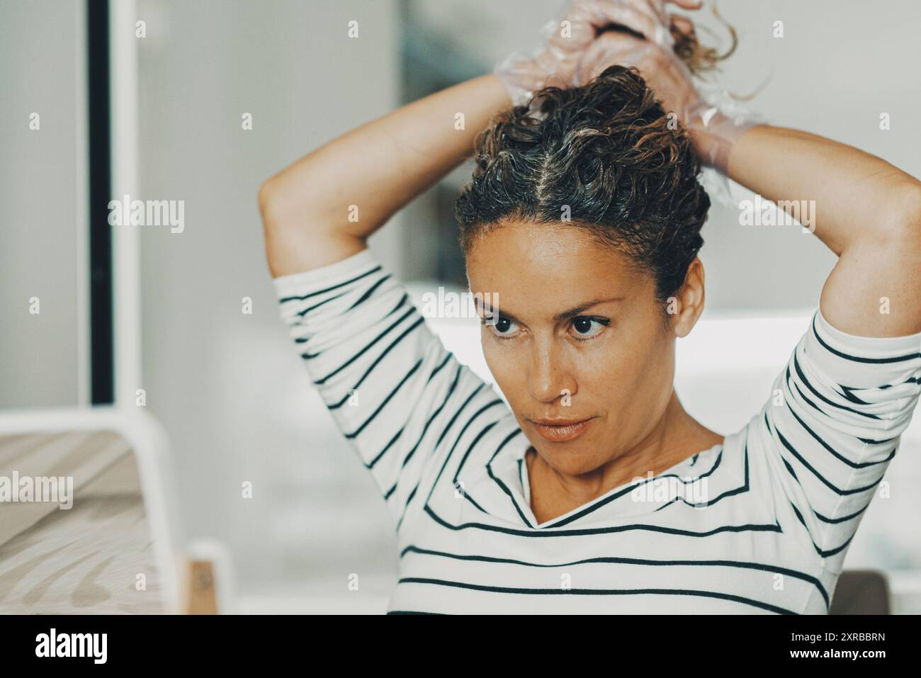 Fare bellezza cura acconciatura persone concetto stile di vita indoor attività di svago a casa. Una giovane e graziosa donna adulta che si fa acconciatura guardando nello specchio in cucina. Industria della moda dei capelli Foto Stock