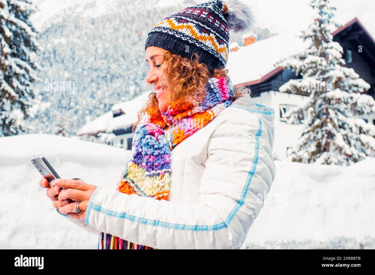 Una donna adulta che usa il telefono in inverno, neve, tempo libero all'aperto, attività tecnologica moderna da sola, sorridente e messaggistica. Vacanze invernali, vacanze estive, donne all'aperto. Cielo blu e sfondo della casa Foto Stock