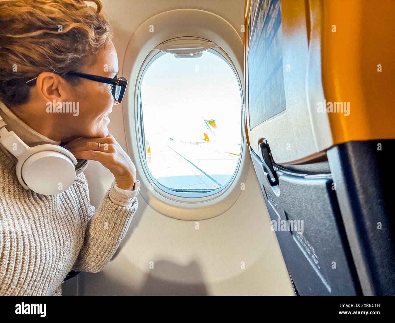 Passeggero sorridente con le cuffie bluetooth che guardano fuori dall'oblò dell'aereo. Donna di mezza età seduta accanto alla finestra, che si rilassa mentre aspetta di arrivare Foto Stock