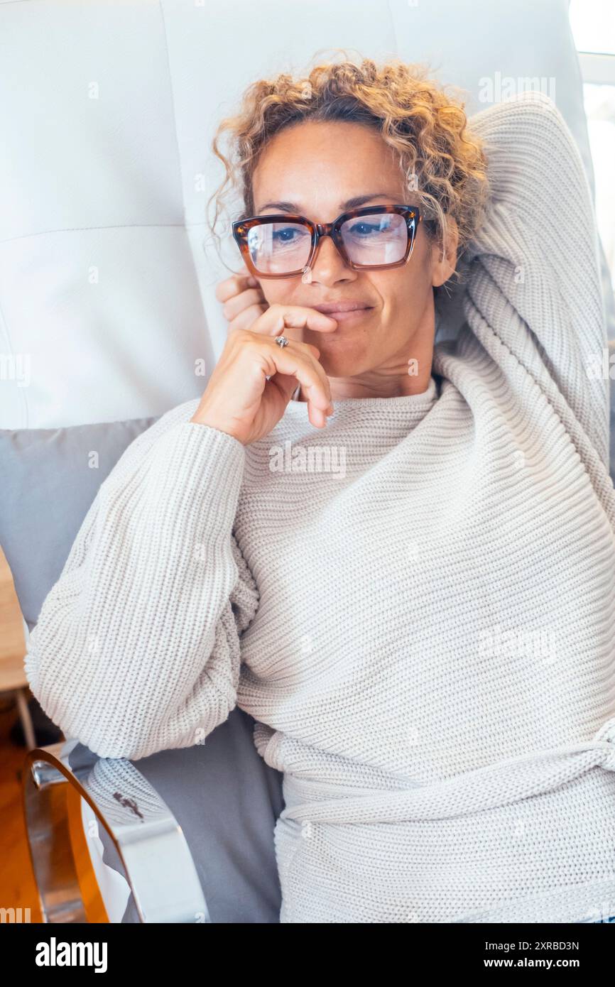 Ritratto di una bella donna matura a casa seduta su un divano che sorride e pensa a uno stile di vita felice. Una donna felice sorride e si diverte a rilassarsi indo Foto Stock