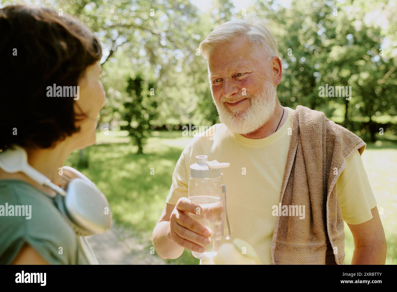 Uomo dai capelli grigi che tiene una bottiglia d'acqua e sorride alla moglie bruna dopo corsa nel parco Foto Stock