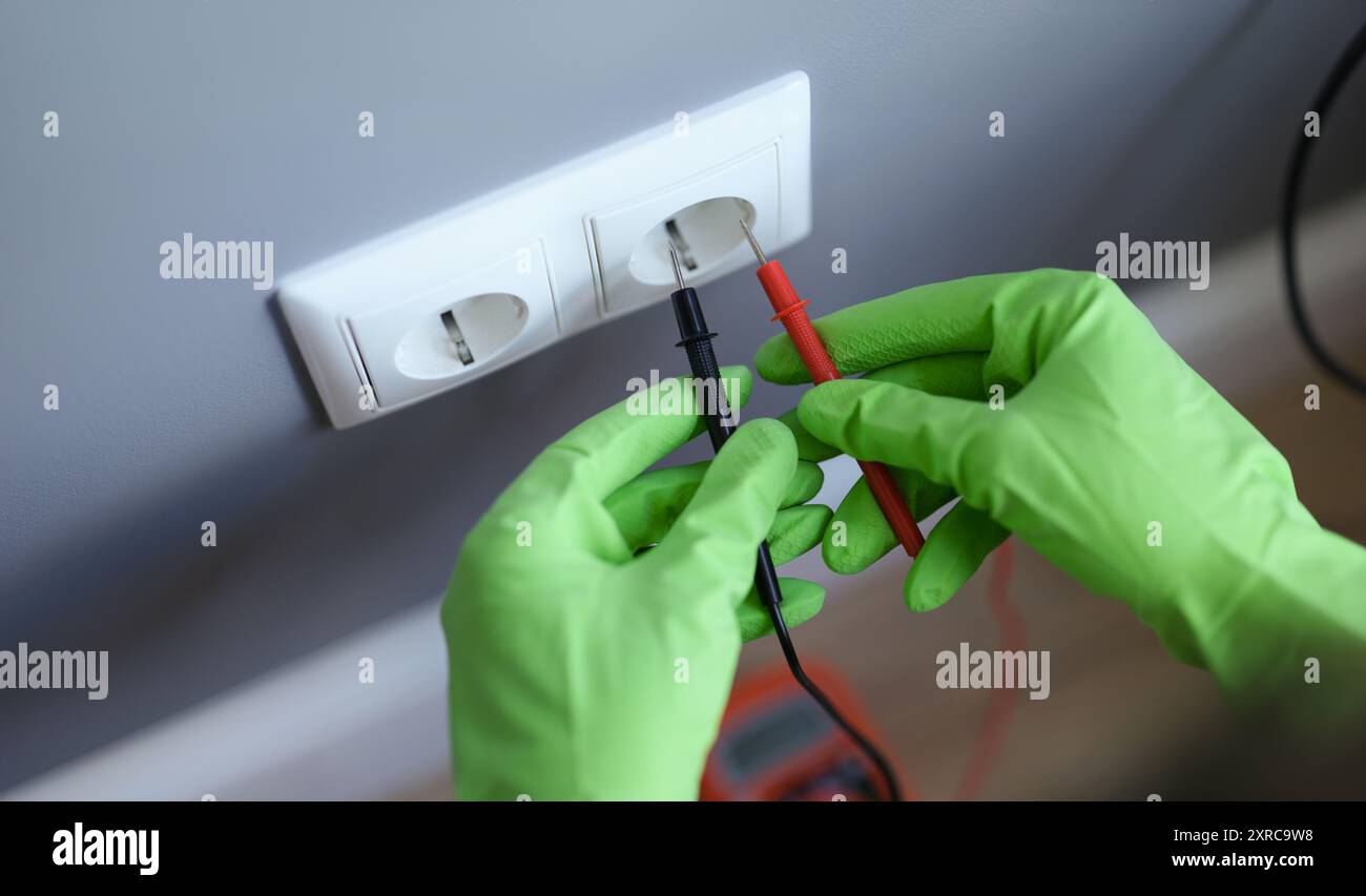 Le mani nei guanti verdi collegano il voltmetro a una presa a muro Foto Stock