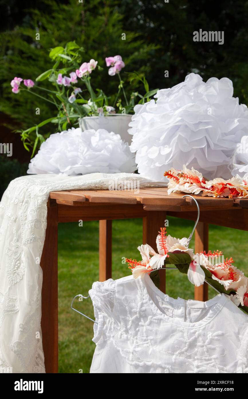 Primo piano di un vestito bianco per bambini con pizzo e una corona di fiori come copricapo appeso su un appendiabiti a tavola, preparativi per un matrimonio, preparazione all'aperto Foto Stock