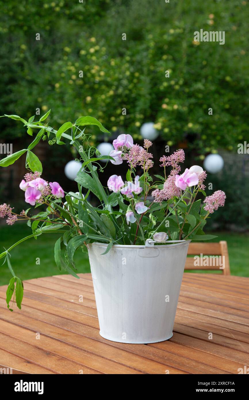 Un cachepot bianco con fiori freschi da giardino come i sacchetti rosa, vetche bianche e rosa e foglie verdi si erge come una disposizione floreale su un tavolo di legno nel giardino; un albero di mele con mele verdi chiare si staglia sfocato sullo sfondo Foto Stock