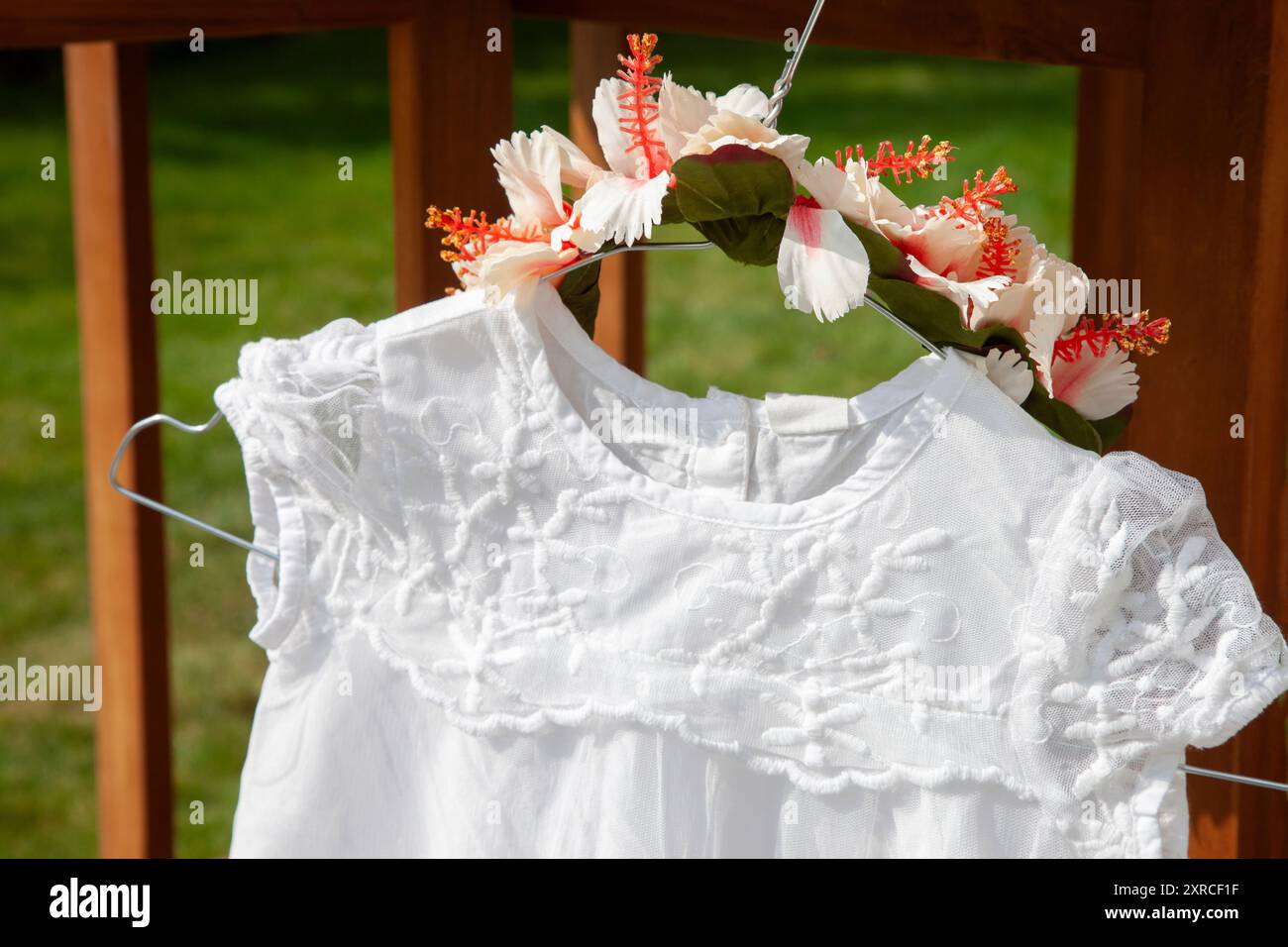 Primo piano di un vestito bianco per bambini con pizzo e una corona di fiori come copricapo appeso a un gancio di filo su un tavolo, preparativi per un matrimonio, preparazione all'aperto Foto Stock