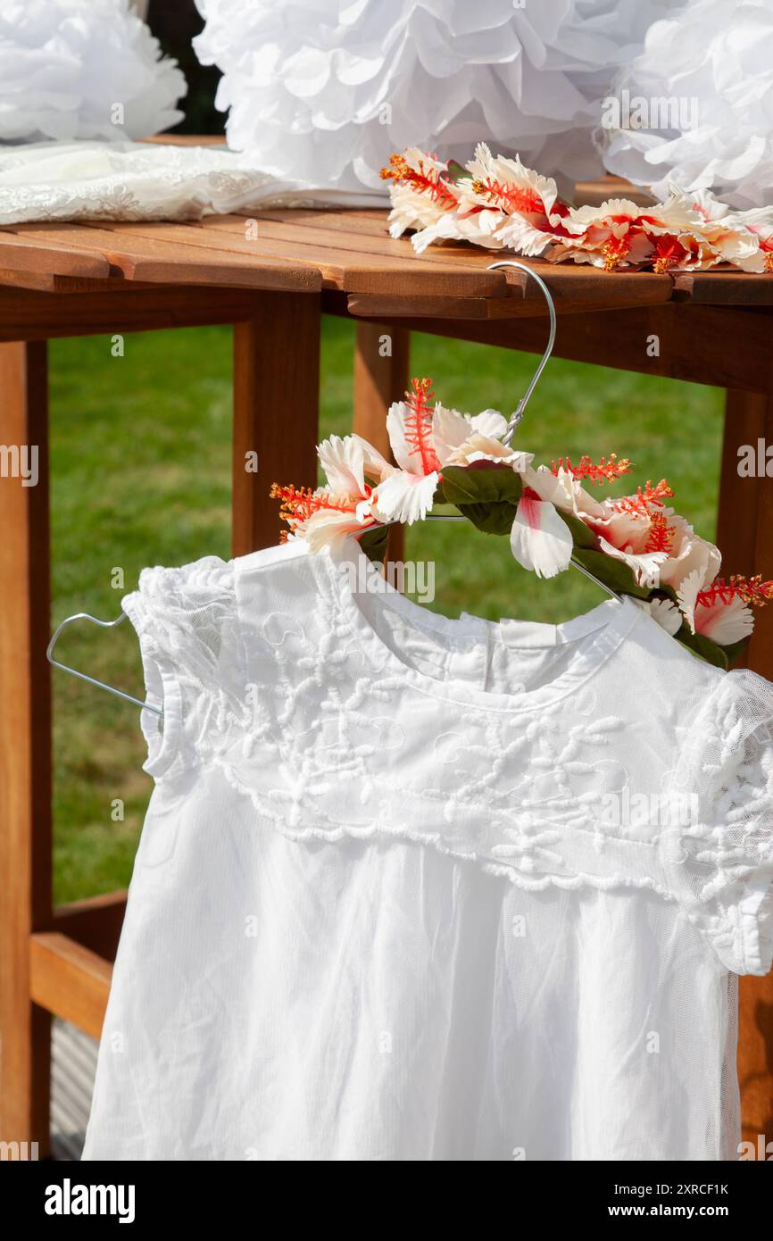 Primo piano di un vestito bianco per bambini con pizzo e una corona di fiori come copricapo appeso su un appendiabiti a un tavolo, preparativi per un matrimonio, preparazione all'aperto Foto Stock