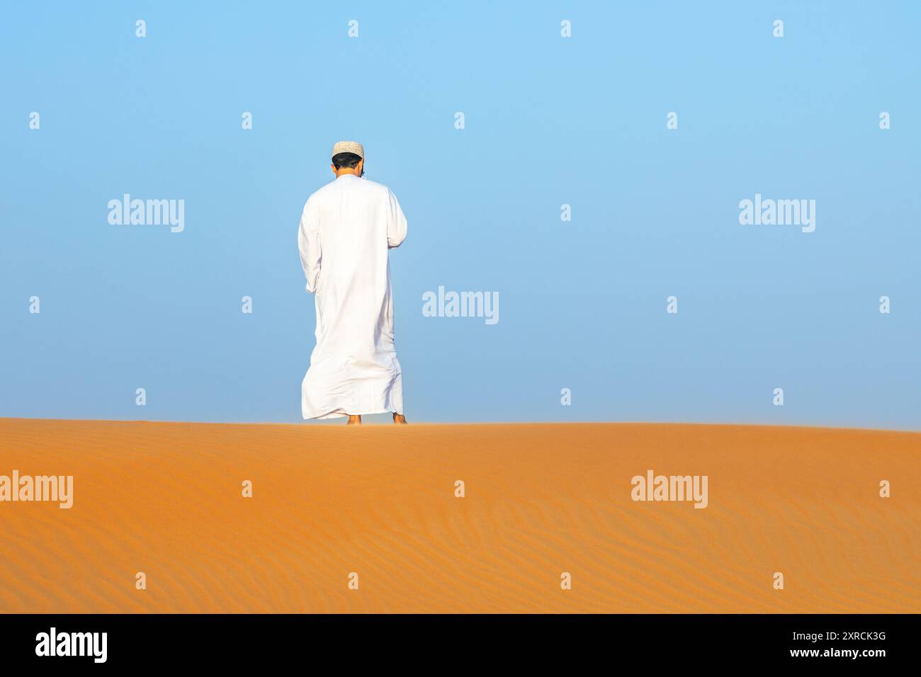 Uomo dell'Oman in abito lungo bianco in piedi sulle dune di sabbia del deserto arancione nel mezzo del deserto di Wahiba, Oman Foto Stock